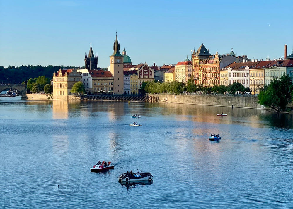 View of paddle boats on the Vltava River with colorful historic buildings and towers in Prague, Czech Republic.